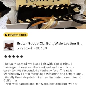 May include: A black suede belt featuring gold metallic lacing and gold-toned tassels, presented in a white box with a cream-colored fabric pouch labeled JUAN.JO. The belt is displayed inside white tissue paper, highlighting the contrast between the black material and the metallic gold accents.