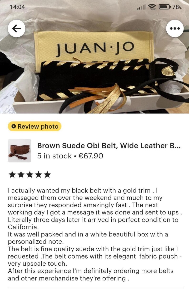 May include: A black suede belt featuring gold metallic lacing and gold-toned tassels, presented in a white box with a cream-colored fabric pouch labeled JUAN.JO. The belt is displayed inside white tissue paper, highlighting the contrast between the black material and the metallic gold accents.
