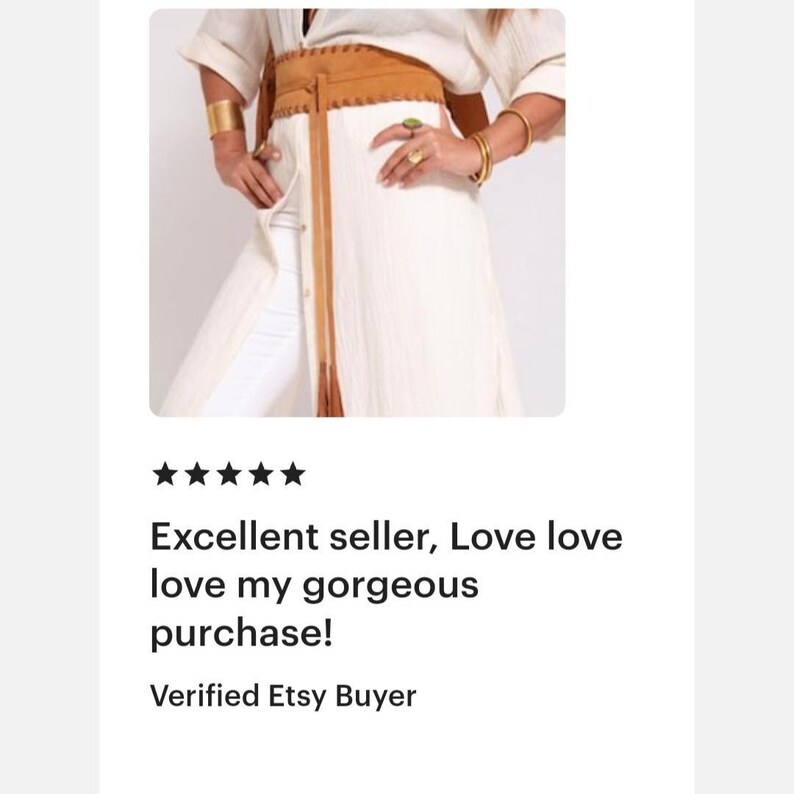May include: A wide tan suede wrap belt with stitched edges and long hanging ties, worn over a long-sleeved white button-down shirt and white pants. The belt features a textured finish and is tied at the waist with two long, thin suede straps.