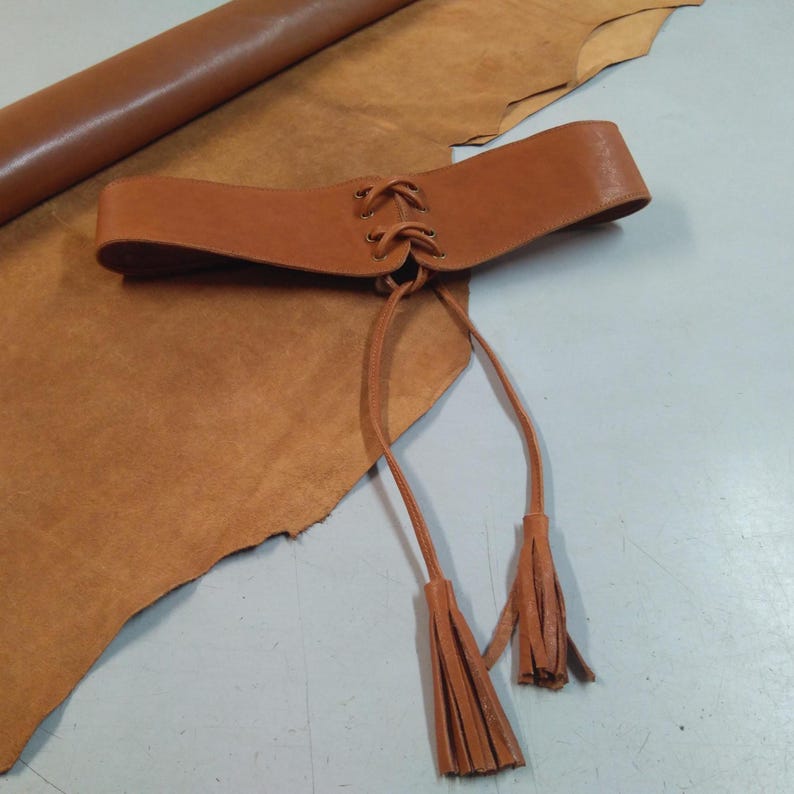 May include: A wide, tan leather belt with a lace-up front and long, tassel-ended ties. The belt is displayed on a piece of tan leather. The belt's width is approximately 4 inches.