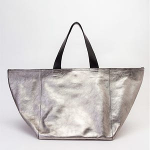 May include: A silver metallic tote bag with black leather handles. The bag has a wide, open top and a spacious interior.