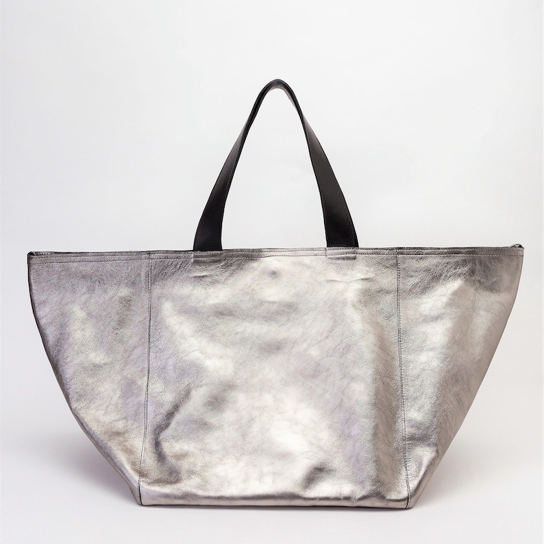 May include: A silver metallic tote bag with black leather handles. The bag has a wide, open top and a spacious interior.