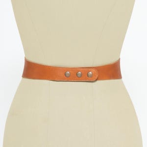 Back view of a handmade tan brown leather corset belt displayed on a mannequin. The belt is secured with three metallic snap closures, ensuring a comfortable and adjustable fit. The smooth, high-quality leather provides structure while allowing flexibility. At the front, it features a lace-up design with metal eyelets and long leather ties ending in tassels, enhancing its bohemian and waist-cinching style