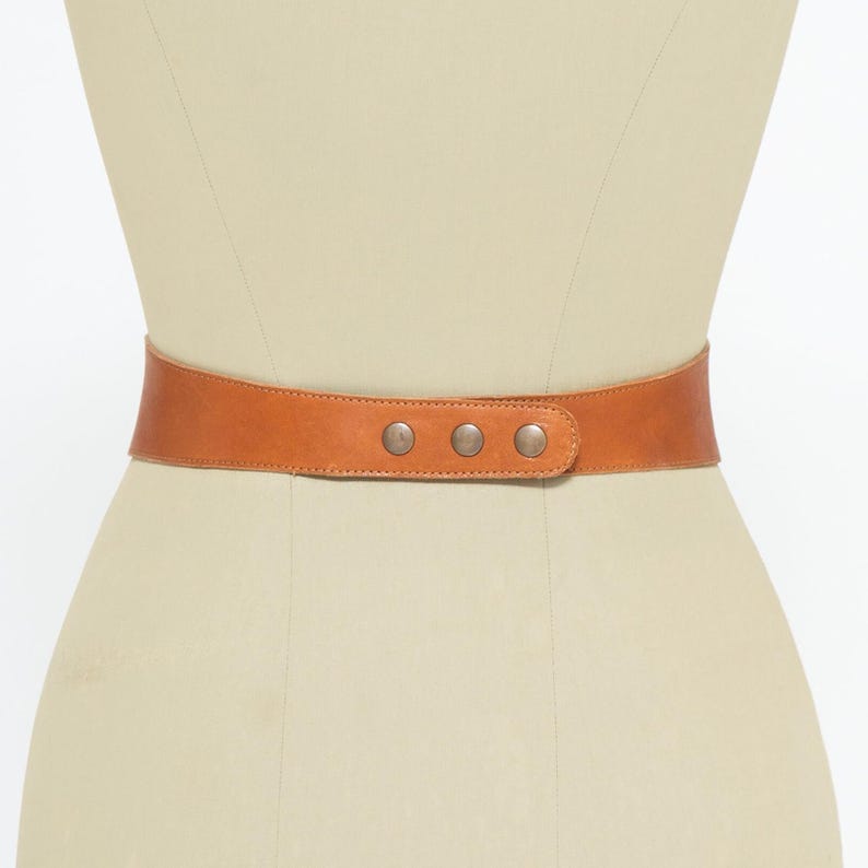 Back view of a handmade tan brown leather corset belt displayed on a mannequin. The belt is secured with three metallic snap closures, ensuring a comfortable and adjustable fit. The smooth, high-quality leather provides structure while allowing flexibility. At the front, it features a lace-up design with metal eyelets and long leather ties ending in tassels, enhancing its bohemian and waist-cinching style