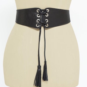 May include: A black corset belt with silver-toned grommets and a lace-up design. The belt features two long, thin ties with matching black tassels at the ends. The belt is displayed on a mannequin.