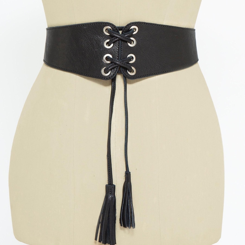 May include: A black corset belt with silver-toned grommets and a lace-up design. The belt features two long, thin ties with matching black tassels at the ends. The belt is displayed on a mannequin.