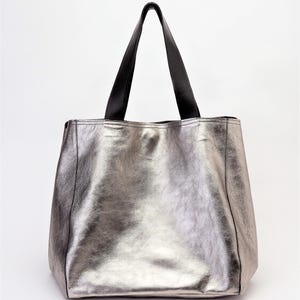 Side view of an extra-large metallic silver leather tote bag. Its shiny and smooth leather reflects light with a sophisticated effect. The reinforced black leather handles add contrast and elegance, allowing for comfortable wear on the shoulder or by hand. The structured design enhances the beauty of the high-quality leather, offering a luxurious and modern finish.