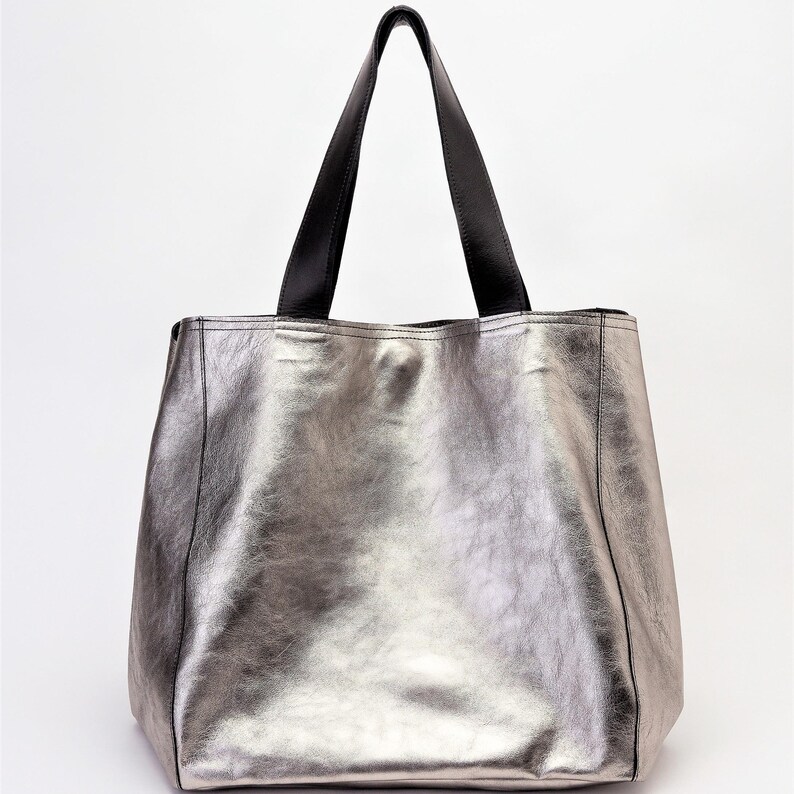 Side view of an extra-large metallic silver leather tote bag. Its shiny and smooth leather reflects light with a sophisticated effect. The reinforced black leather handles add contrast and elegance, allowing for comfortable wear on the shoulder or by hand. The structured design enhances the beauty of the high-quality leather, offering a luxurious and modern finish.