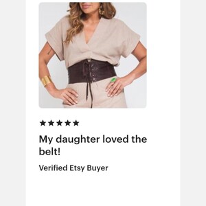 Op de afbeelding: Een brede, donkerbruine korset-stijl riem met een vetersluiting aan de voorzijde en bijpassende bruine veters. De riem wordt gedragen over een lichtbeige top met korte mouwen, een V-hals en omgeslagen mouwen voor een gestructureerd silhouet.