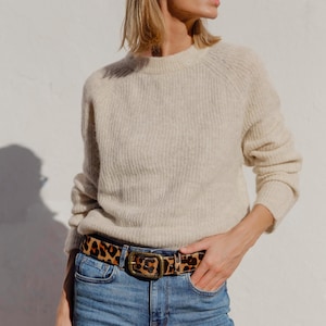 Könnte beinhalten: Ein beigefarbener, langärmeliger Strickpullover mit gerippter Textur und Rundhalsausschnitt, kombiniert mit einer blauen Jeans und einem Gürtel mit braun-schwarzem Leopardenmuster sowie einer rechteckigen, messingfarbenen Metallschnalle.