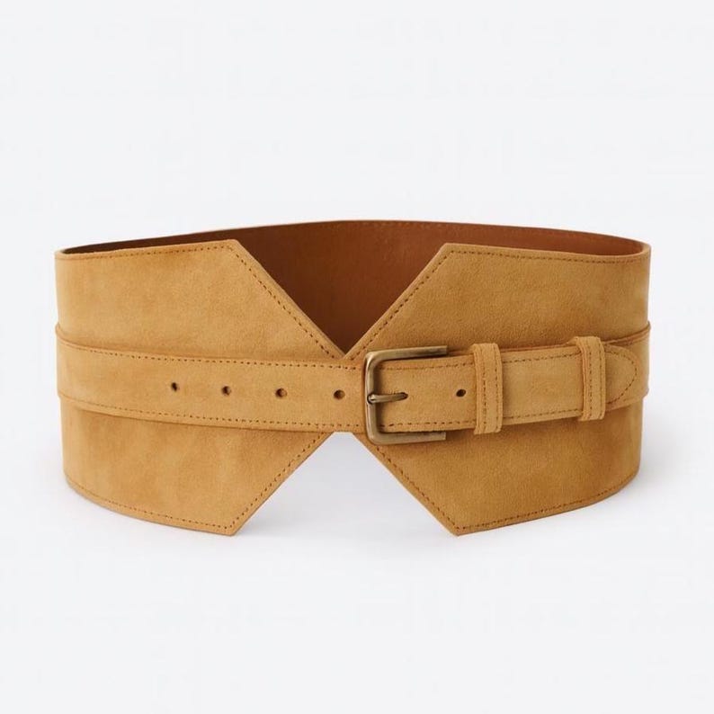May include: A wide tan suede belt featuring a structured, angular design that tapers toward the center. The belt includes a matching tan strap with a series of punched holes and a rectangular brass-toned metal buckle for secure fastening.