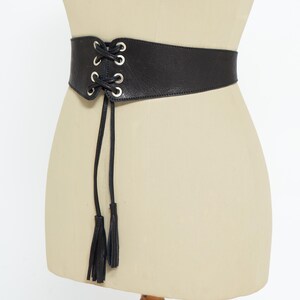 May include: Black leather corset belt with silver-tone grommets and a lace-up front. The belt features two long black tassels hanging from the front. The belt is displayed on a mannequin.