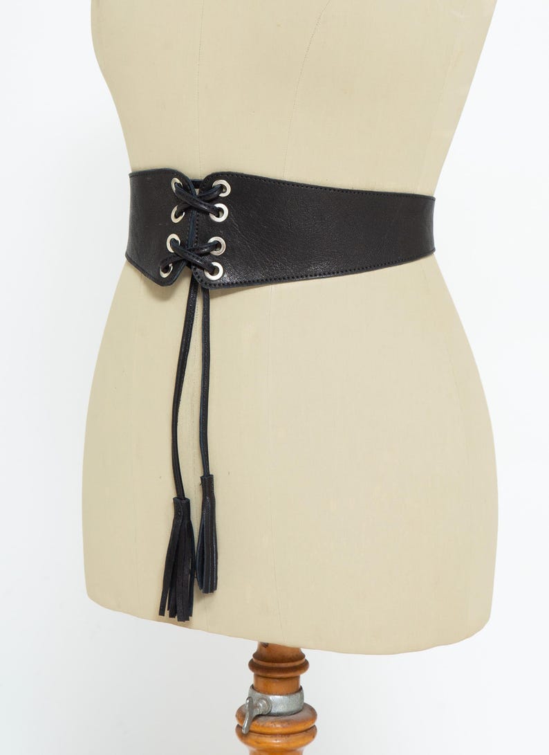 May include: Black leather corset belt with silver-tone grommets and a lace-up front. The belt features two long black tassels hanging from the front. The belt is displayed on a mannequin.