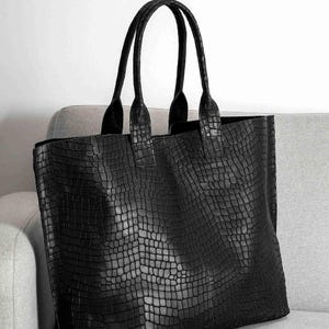 May include: A black tote bag featuring a textured crocodile skin pattern across the entire surface. The bag has two tall, matching black handles attached to the top edge, providing a structured rectangular shape with a consistent dark finish.