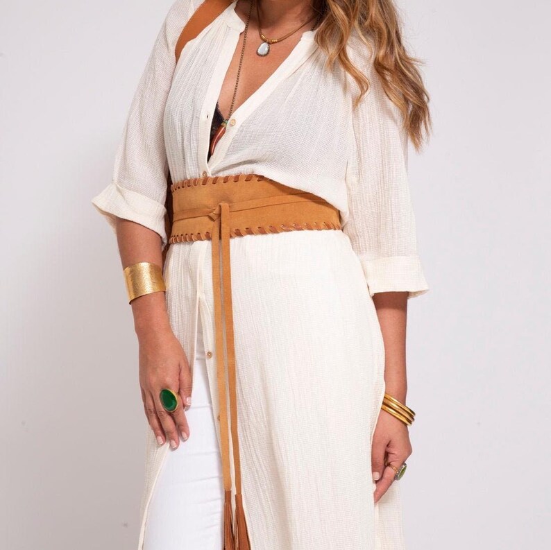 May include: A tan suede wrap belt with a lace-up front detail worn over a long-sleeved white button-down shirt and white pants. The outfit is accessorized with a gold cuff bracelet, a gold bangle set, and a ring featuring a large green stone.