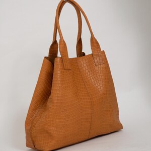 May include: A tan tote bag featuring a textured crocodile-embossed pattern across the entire exterior. The bag has two tall, matching tan handles attached to the top, a vertical seam running down the center, and an open-top design.