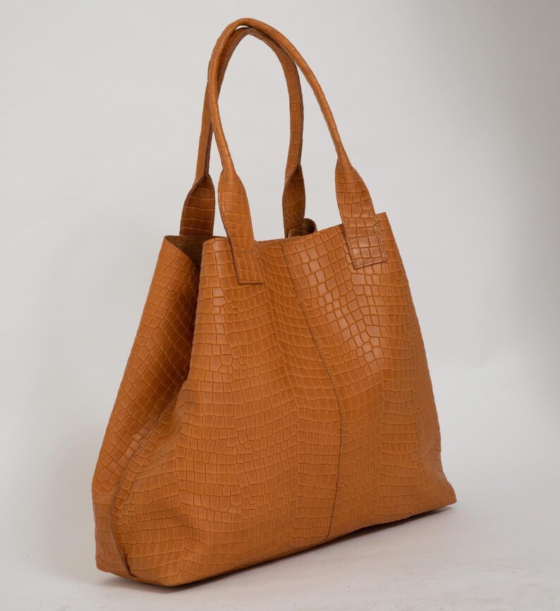 May include: A tan tote bag featuring a textured crocodile-embossed pattern across the entire exterior. The bag has two tall, matching tan handles attached to the top, a vertical seam running down the center, and an open-top design.