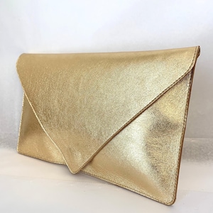 Peut inclure: Une pochette enveloppe dorée métallisée avec une fermeture à rabat triangulaire. L'extérieur présente une finition texturée et scintillante avec des coutures visibles le long des bords, offrant une silhouette rectangulaire pour transporter des objets.