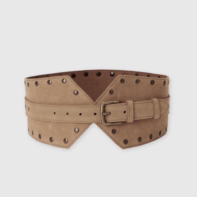 May include: A wide tan suede belt featuring a central strap with a metal buckle and a series of evenly spaced metal studs along the top and bottom edges. The belt has a distinct angular shape that tapers toward the center buckle closure.