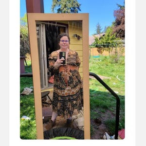 May include: A full-length mirror reflects a person wearing a long, patterned dress with a brown corset belt. The dress features a black background with floral and paisley designs in shades of orange and beige. The mirror has a light brown wooden frame.