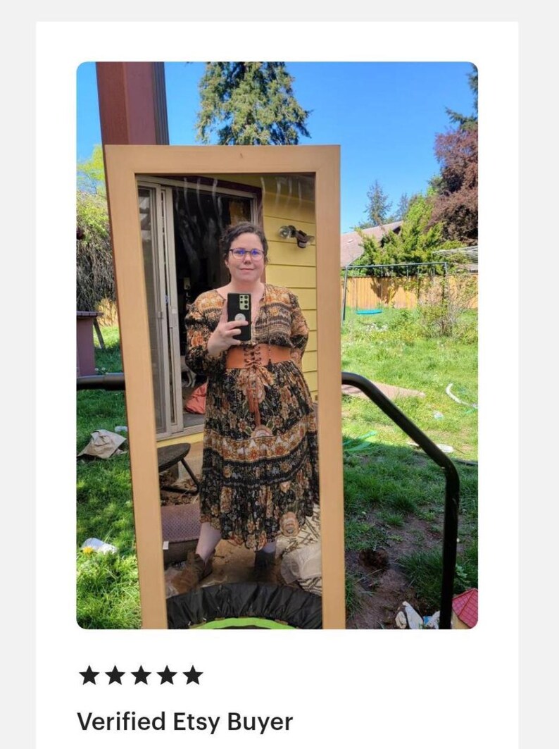 May include: A full-length mirror reflects a person wearing a long, patterned dress with a brown corset belt. The dress features a black background with floral and paisley designs in shades of orange and beige. The mirror has a light brown wooden frame.