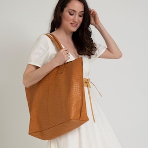 May include: A large, tan-colored tote bag featuring a textured, embossed crocodile-skin pattern. The bag has two sturdy shoulder straps and a spacious, rectangular silhouette. The material has a matte finish and a consistent, uniform tan color.