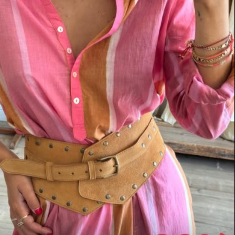May include: A long-sleeved button-down shirt featuring vertical stripes in shades of pink, white, and tan. A wide, tan suede corset-style belt with a metal buckle and silver-toned studs is worn over the shirt. Several thin bracelets are on the wrist.