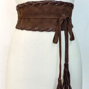 May include: A wide dark brown waist belt featuring a smooth central panel and top and bottom edges finished with a decorative dark brown leather whipstitch lacing. The belt secures with long thin brown ties that hang down with tassel ends.