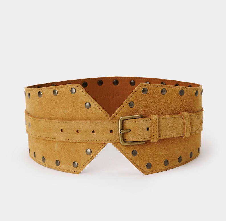 May include: A wide mustard yellow suede belt featuring a central buckle closure and a series of metallic studs along the top and bottom edges. The belt has a structured, contoured shape with a horizontal strap overlay across the middle section.