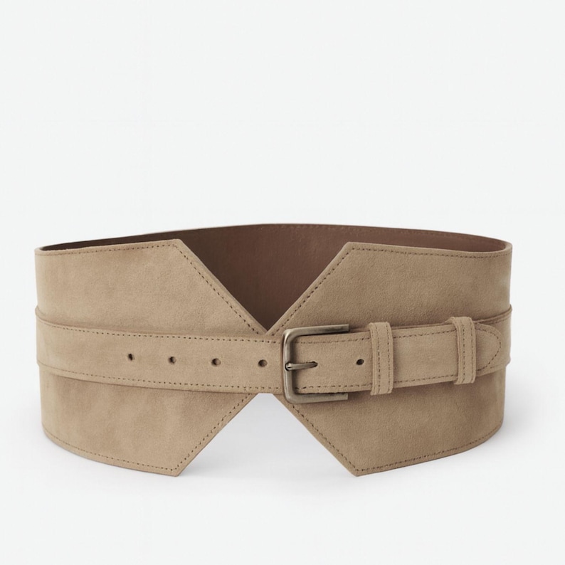 May include: A wide, beige, suede-like waist belt featuring a structured, angular front design with a pointed V-shape cutout. The belt includes a matching horizontal strap across the center, secured by a rectangular, antique-toned metal buckle.