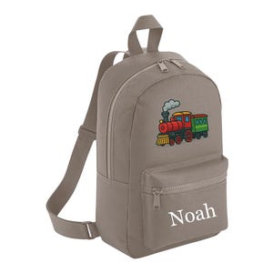May include: A taupe-coloured backpack with a front pocket and a top handle. The backpack features a colourful embroidered train design and the name "Noah" in white. The bag has adjustable straps and a zip closure.