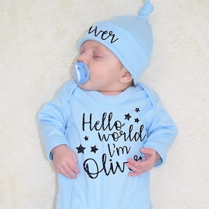 May include: A light blue baby onesie with the text "Hello world I'm Oliver" in black with glitter stars. The baby is wearing a matching light blue hat with the text "Oliver" in black.