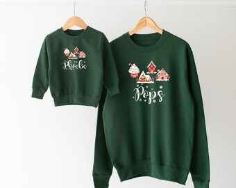May include: Two green Christmas sweaters with gingerbread house designs. The smaller sweater says "Phoebe" and the larger sweater says "Pops".