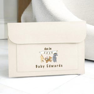May include: Cream-coloured baby announcement clutch with a flap closure. The front features a Winnie the Pooh design with the text "due in 2025" and "Baby Edwards". The clutch is made of a soft, textured material.