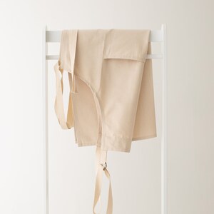 May include: A beige cotton apron with adjustable straps hanging on a white rack.