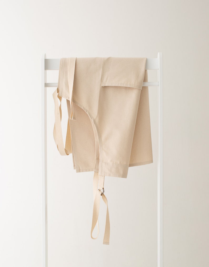 May include: A beige cotton apron with adjustable straps hanging on a white rack.