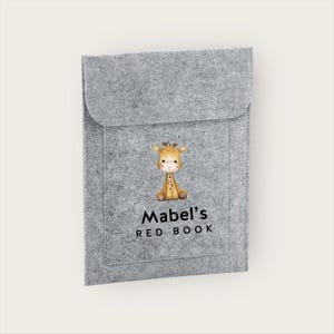 May include: A gray felt book cover with a flap closure. The cover features a watercolor illustration of a giraffe and the text "Mabel's RED BOOK" in black. The book cover is designed to protect a book.