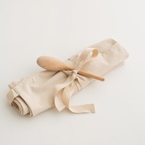 May include: A beige canvas apron rolled up and tied with a beige ribbon, with a wooden spoon placed on top.