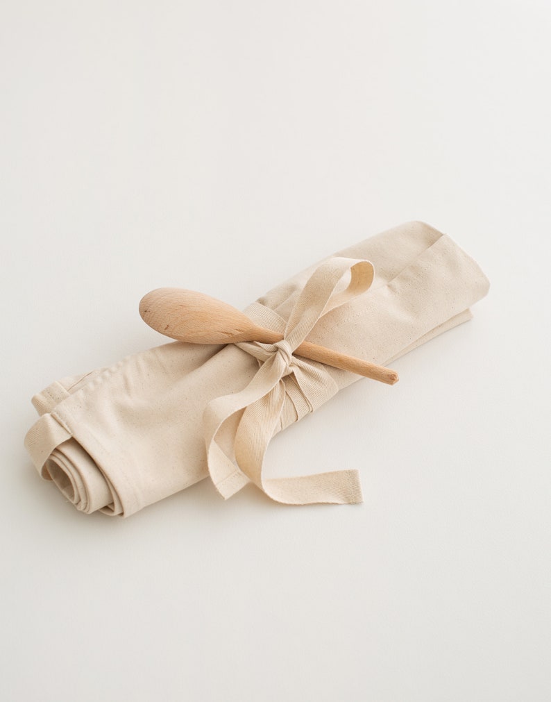 May include: A beige canvas apron rolled up and tied with a beige ribbon, with a wooden spoon placed on top.