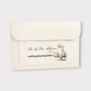 Peut inclure: Un classeur en tissu blanc avec une étiquette qui dit "Mr. & Mrs. Aslam to be wedding planning folder". L'étiquette présente également une illustration aquarelle d'un gâteau de mariage blanc avec des fleurs.