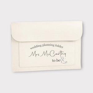 May include: A beige wedding planning folder with the text "wedding planning folder" and "Mrs. McCarthy to be" with a drawing of a white high heel shoe.