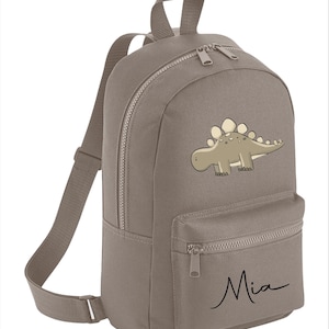 May include: A grey canvas backpack with a cartoon dinosaur design and the name "Mia" embroidered on the front pocket.