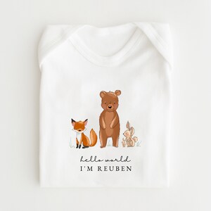 May include: White baby bodysuit with a fox, bear, and rabbit illustration. The text "hello world I'M REUBEN" is printed below the animals. The bodysuit is made of soft material and is designed for newborns.