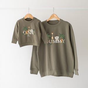 May include: Two olive green sweatshirts hanging on wooden hangers. The smaller sweatshirt says "ONE Theo" with a giraffe, lion, and palm trees. The larger sweatshirt says "MUMMY" with similar animal and plant designs.
