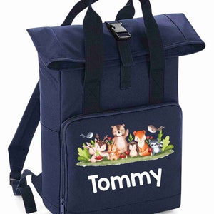 May include: A navy blue canvas backpack with a roll-top closure and a front zippered pocket. The backpack features a colourful woodland animal print with the name "Tommy" printed in white below the animals.