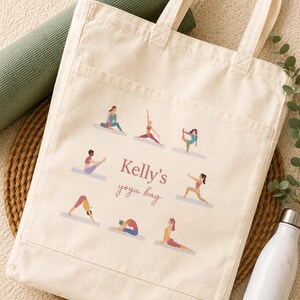 May include: A cream-colored canvas tote bag featuring a front pocket and a printed design of eight figures in various yoga poses surrounding the text "Kelly's yoga bag" in a mix of serif and script fonts in a muted mauve color.