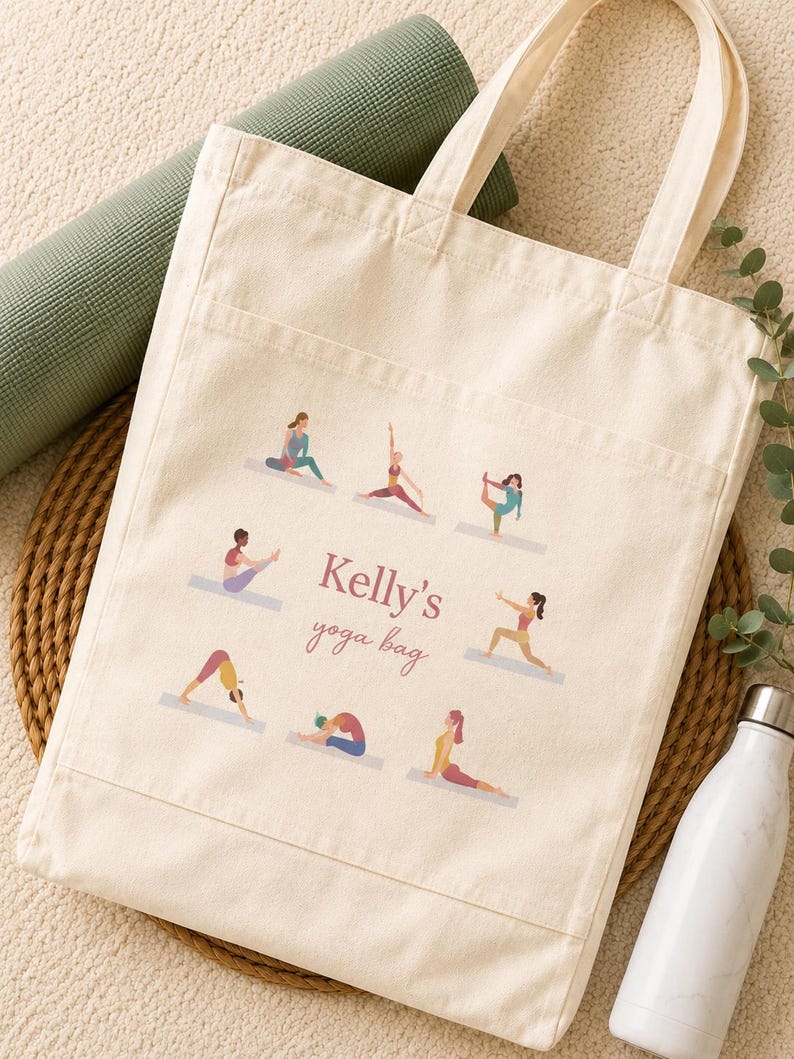 May include: A cream-colored canvas tote bag featuring a front pocket and a printed design of eight figures in various yoga poses surrounding the text "Kelly's yoga bag" in a mix of serif and script fonts in a muted mauve color.