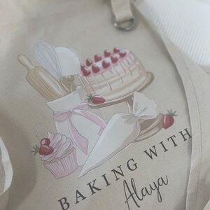 May include: A beige canvas apron with a pink and white cake, cupcake, piping bag, rolling pin, and whisk design. The text "Baking with Alaya" is printed on the apron.