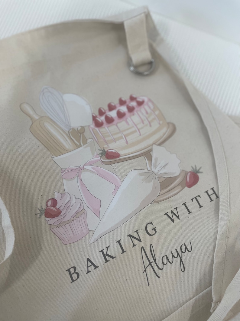May include: A beige canvas apron with a pink and white cake, cupcake, piping bag, rolling pin, and whisk design. The text "Baking with Alaya" is printed on the apron.