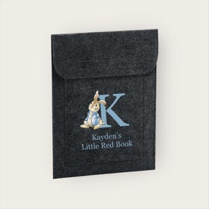 May include: A dark grey felt book cover with a flap. It features a blue letter 'K' and a rabbit illustration in a blue jacket. The text 'Kayden's Little Red Book' is printed below the illustration, suitable for a child's book.